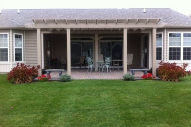 khaki colored pergola attached to a house and over a brick patio area facing the backyard